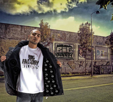 Photographie de DJ Idem posant devant un graffiti Big Factory.